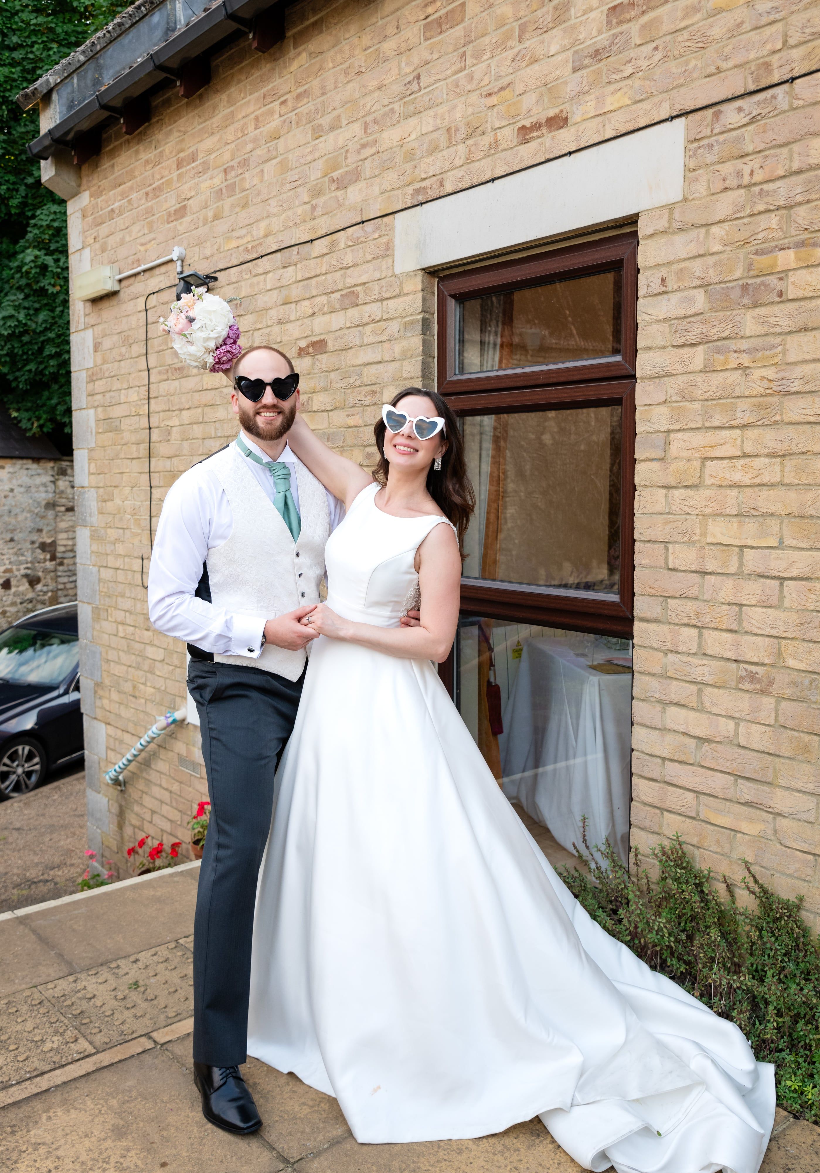 Couple pose in heart sunglasses, bouquet held aloft outside the reception venue