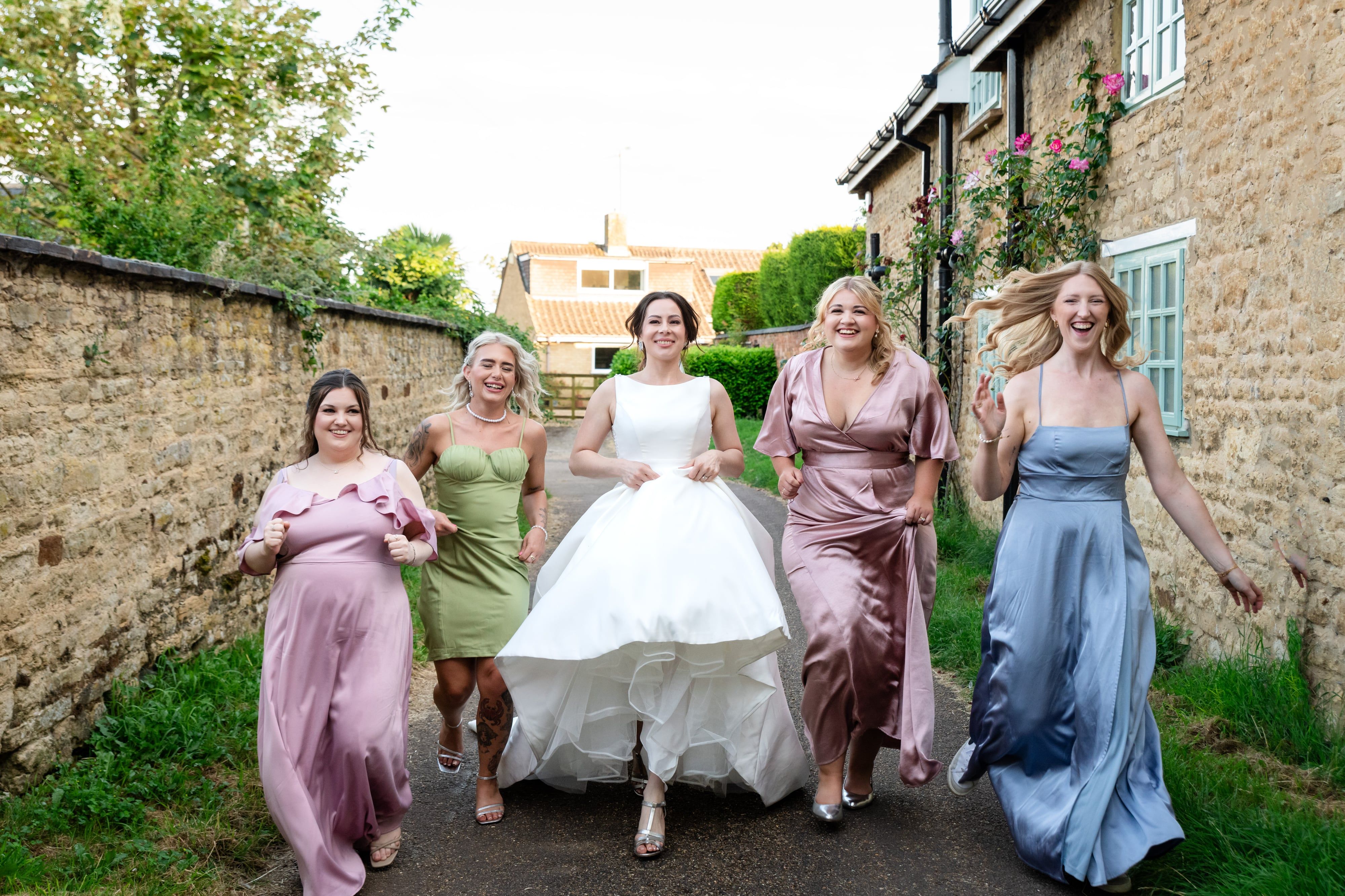 Bride and bridesmaids in mixed pastels walk a stone-walled village lane