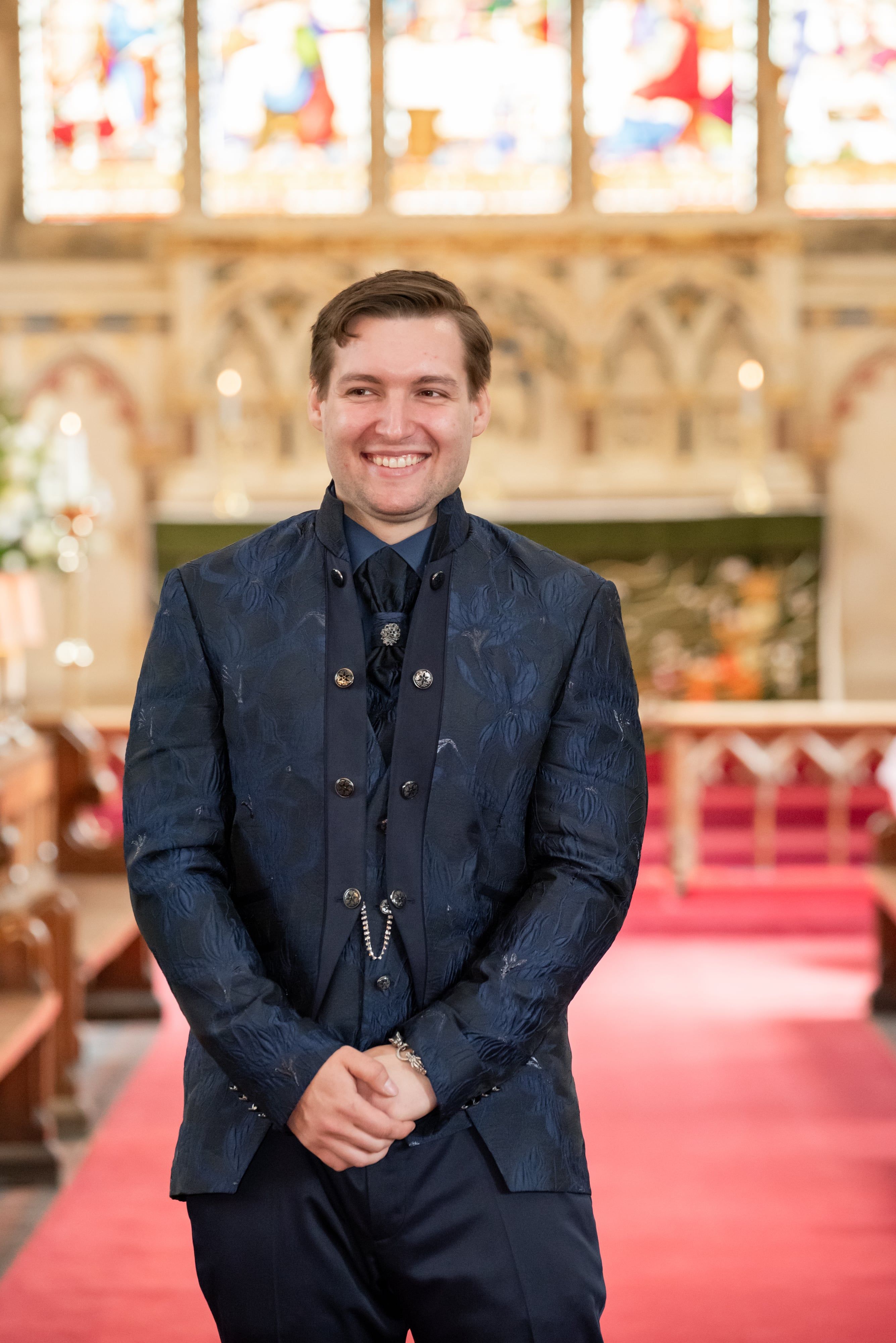 Groom in an embroidered navy jacket smiles at the altar while waiting for the bride