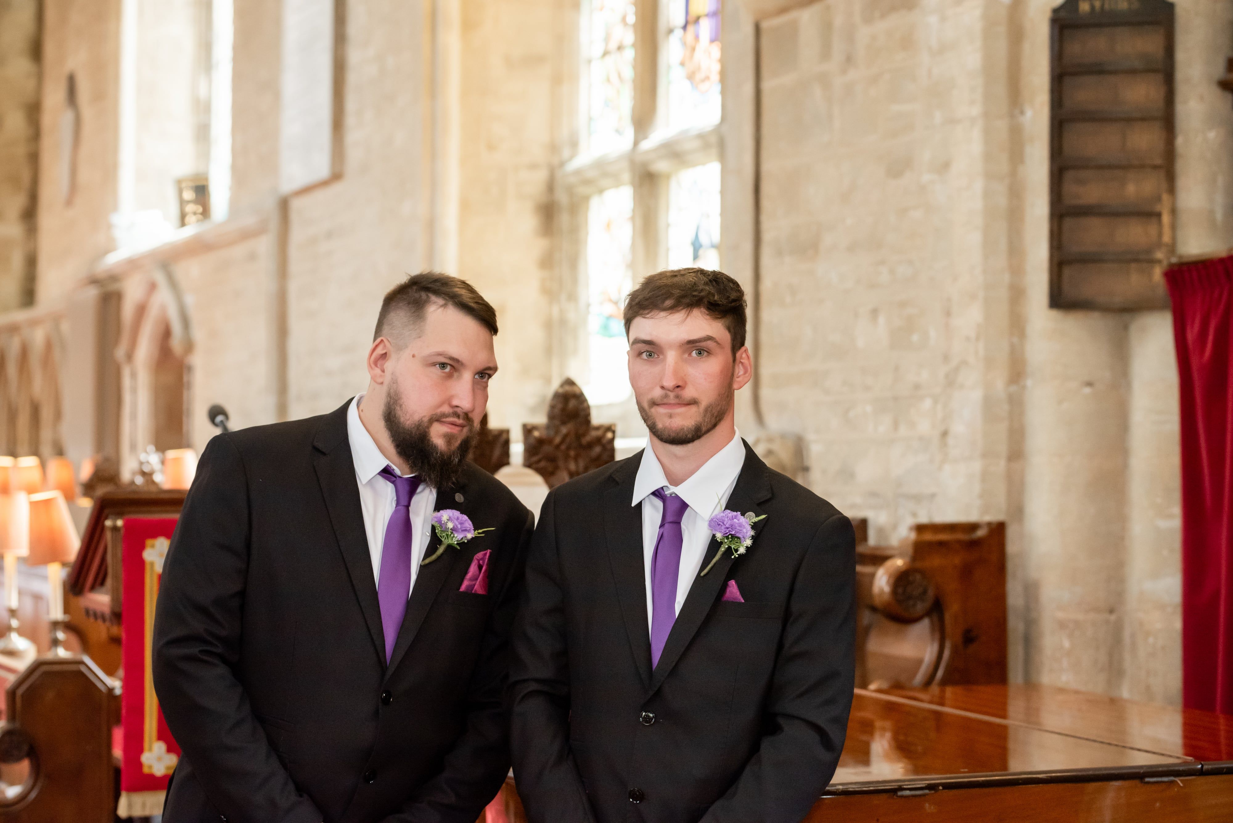 Two best men with purple ties and buttonholes wait by the piano in the church