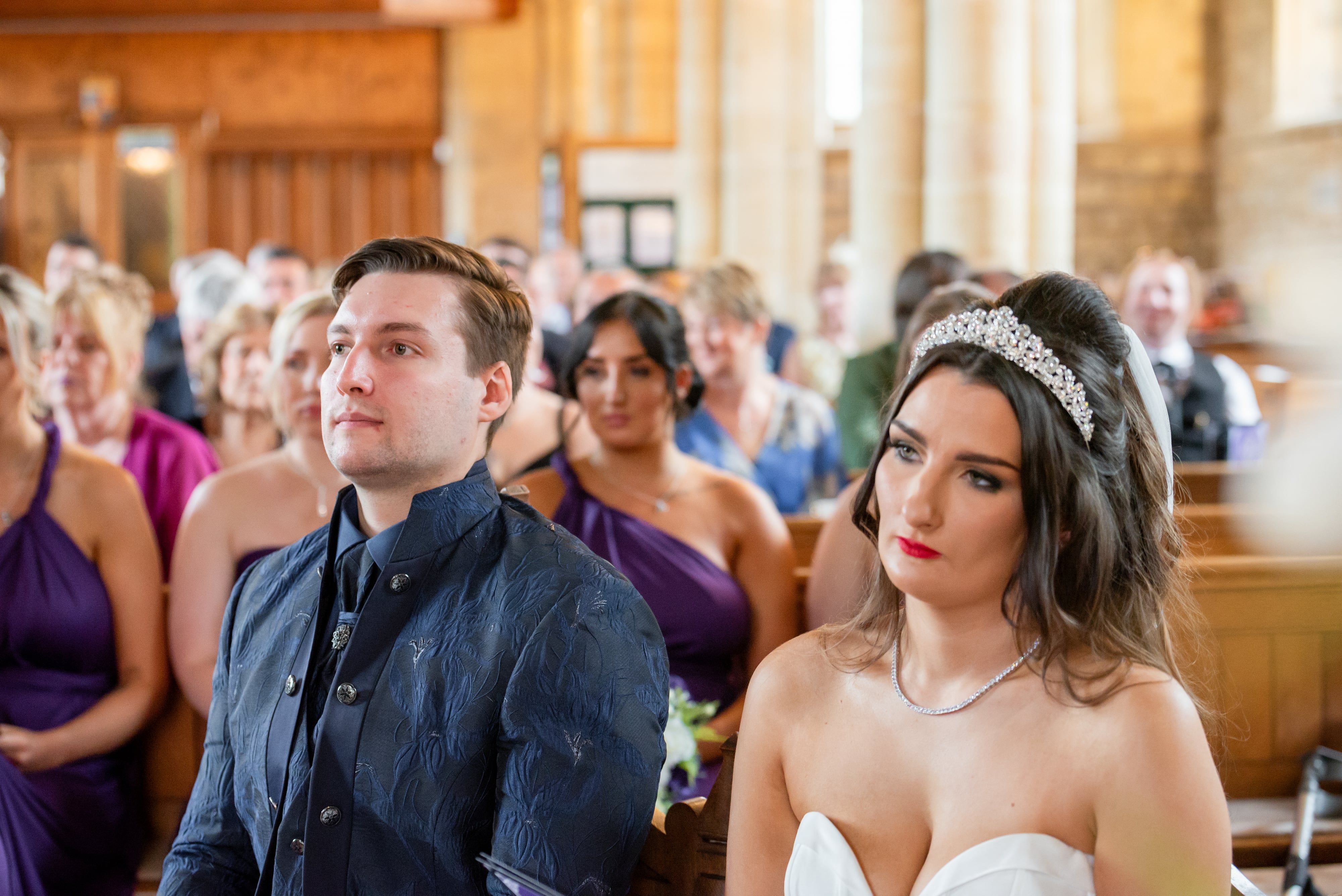 Bride and groom seated side by side during the ceremony, stained glass behind