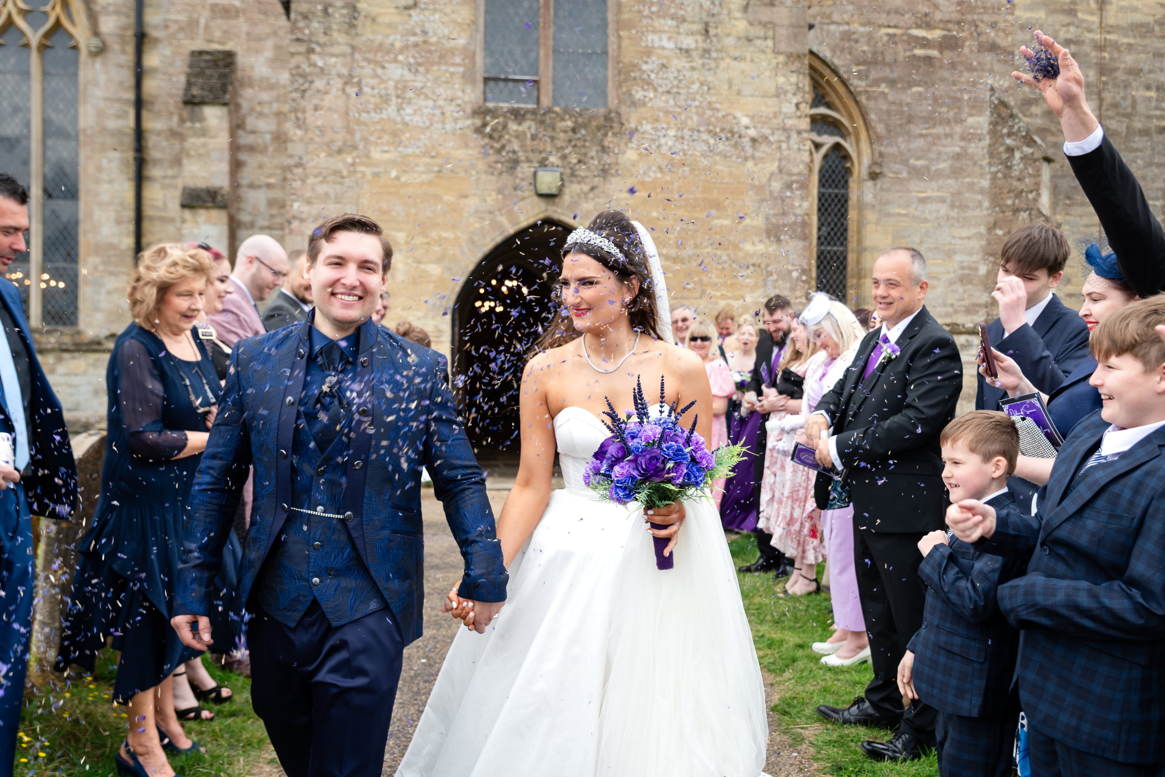 Bride and groom kiss in a confetti shower outside the stone porch of St Peter and St Paul church in Olney