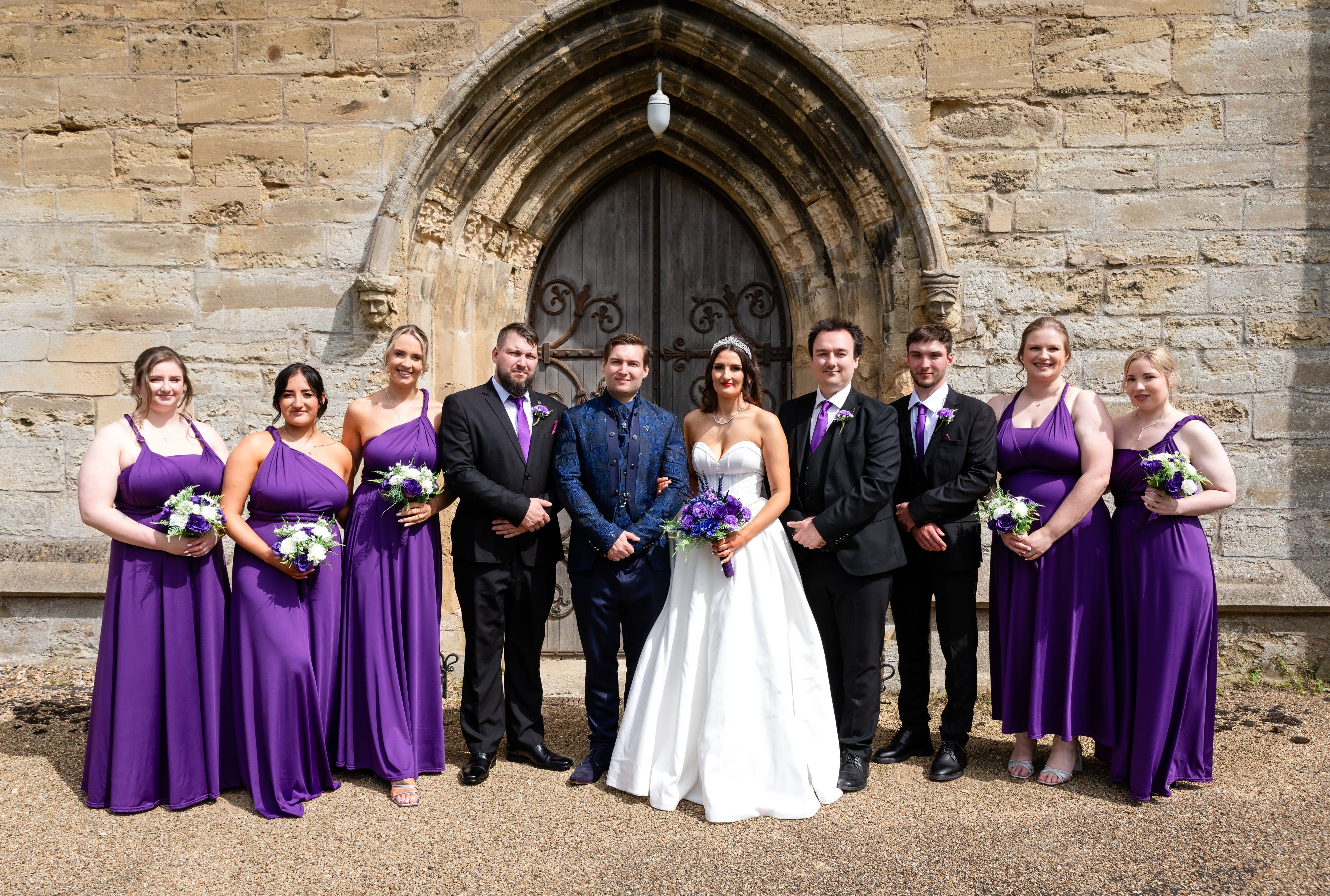 Full bridal party in purple gathered at the arched wooden door of the church