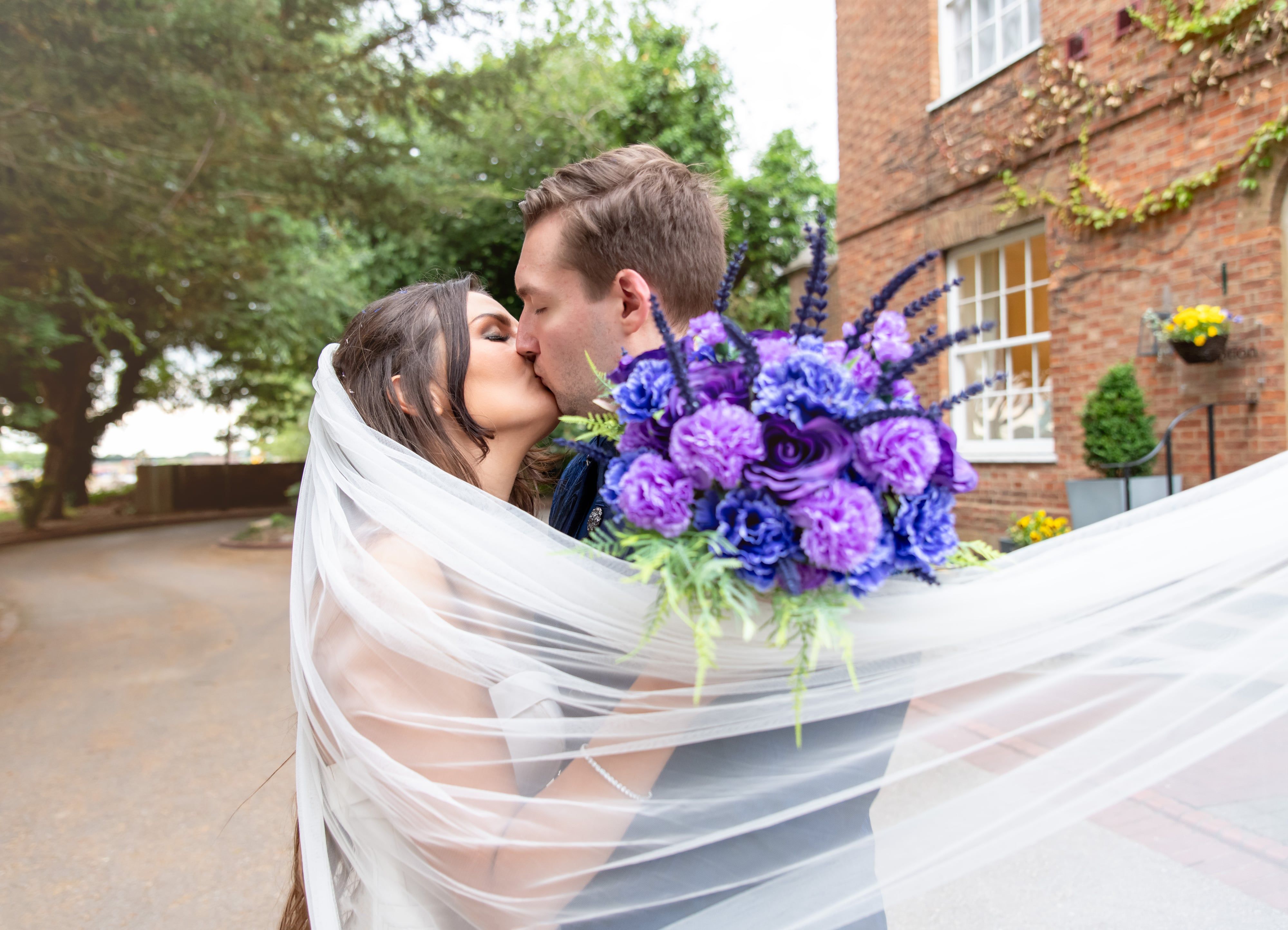 Veil-wrapped kiss with the purple bouquet outside the ivy-clad Georgian frontage of Holiday Inn Milton Keynes East