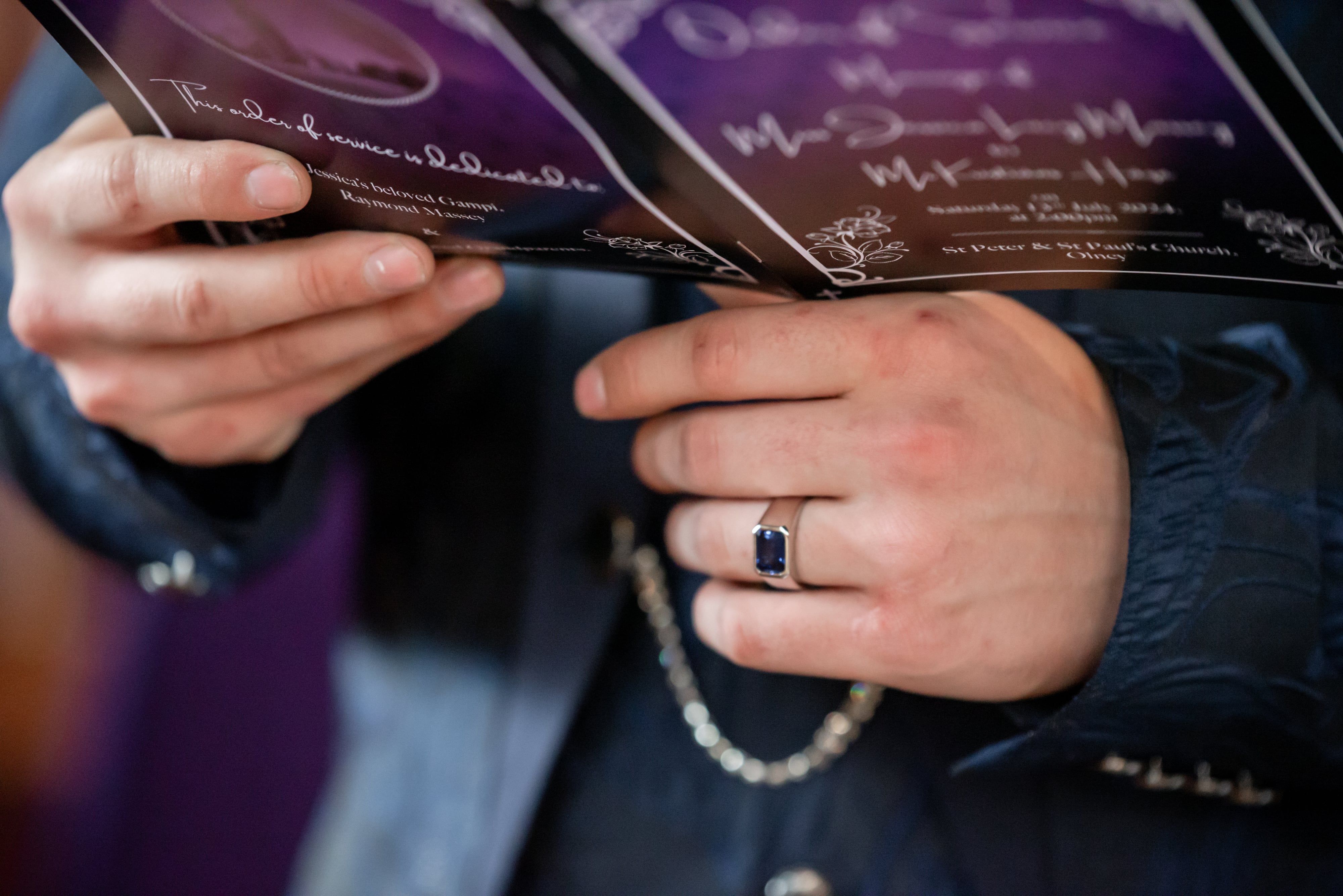 Close-up of the groom's hands holding the order of service, sapphire ring showing