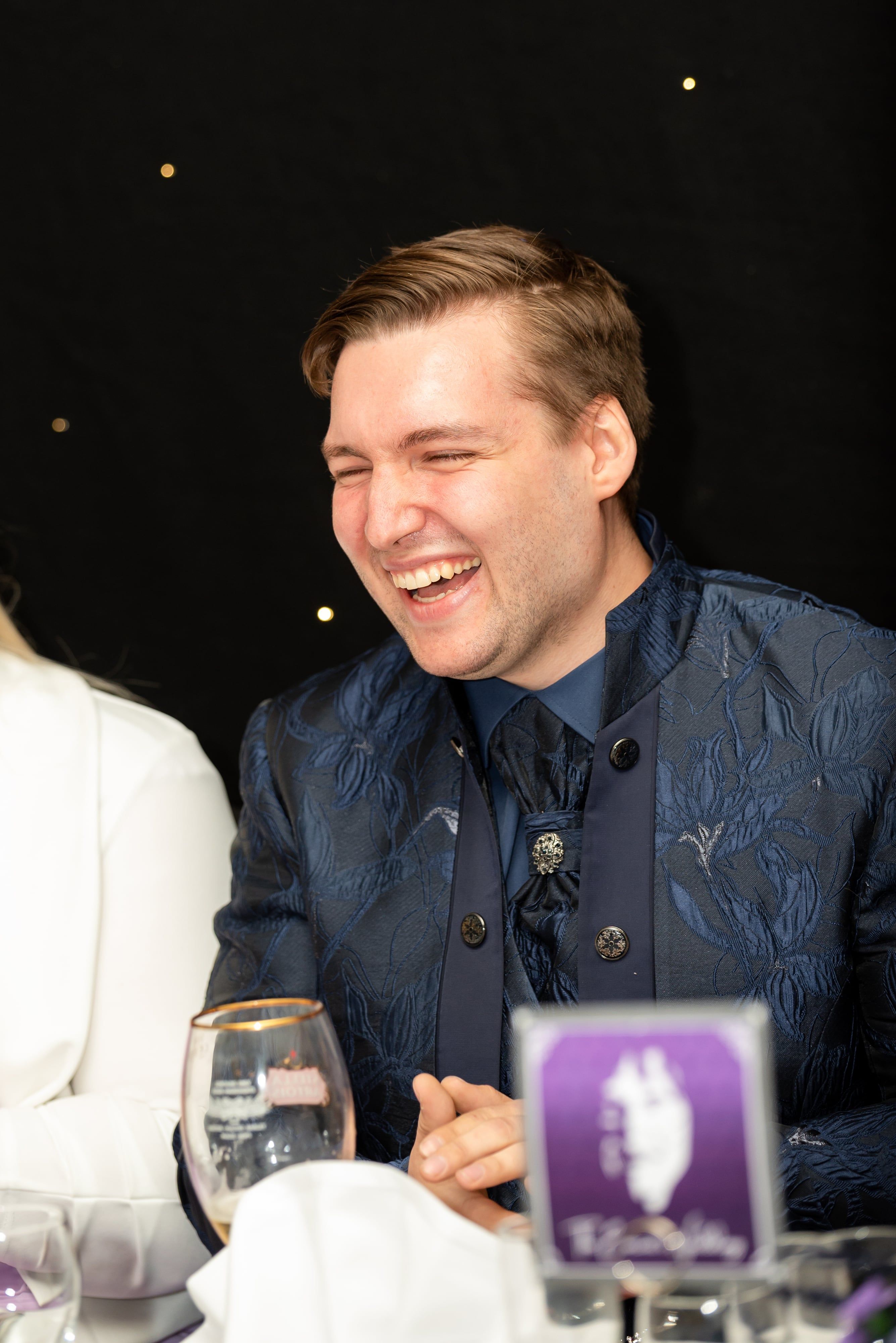 Groom in embroidered navy jacket laughs at the reception table